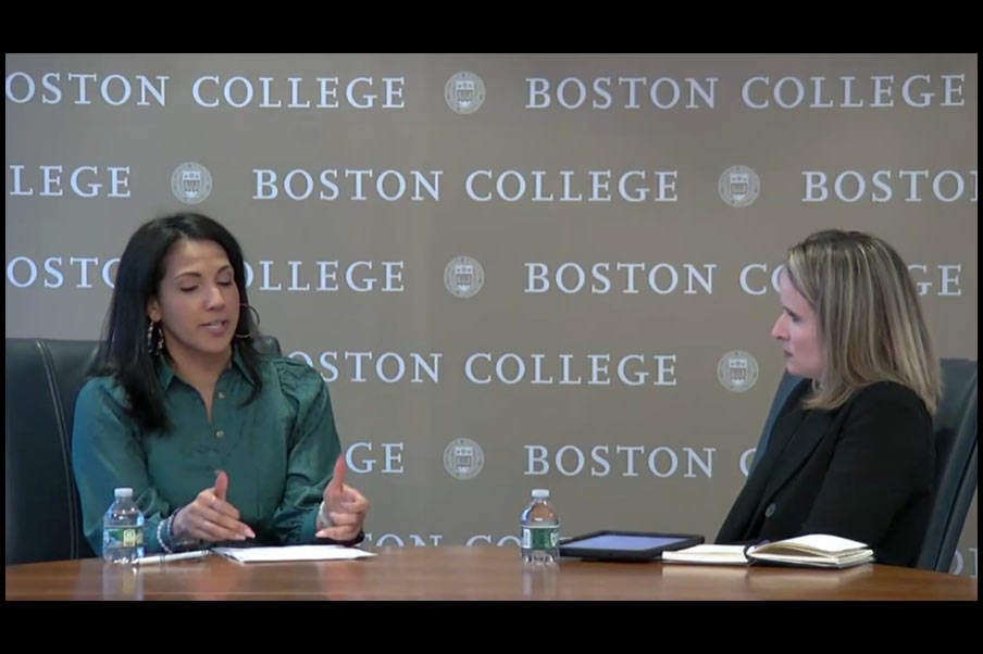 Two women sit at a table having a conversation in front of a Boston College backdrop. One woman gestures with her hands while speaking, and the other listens attentively. Bottled water, a notebook, and a tablet with the DEV_Home24 app open are on the table.