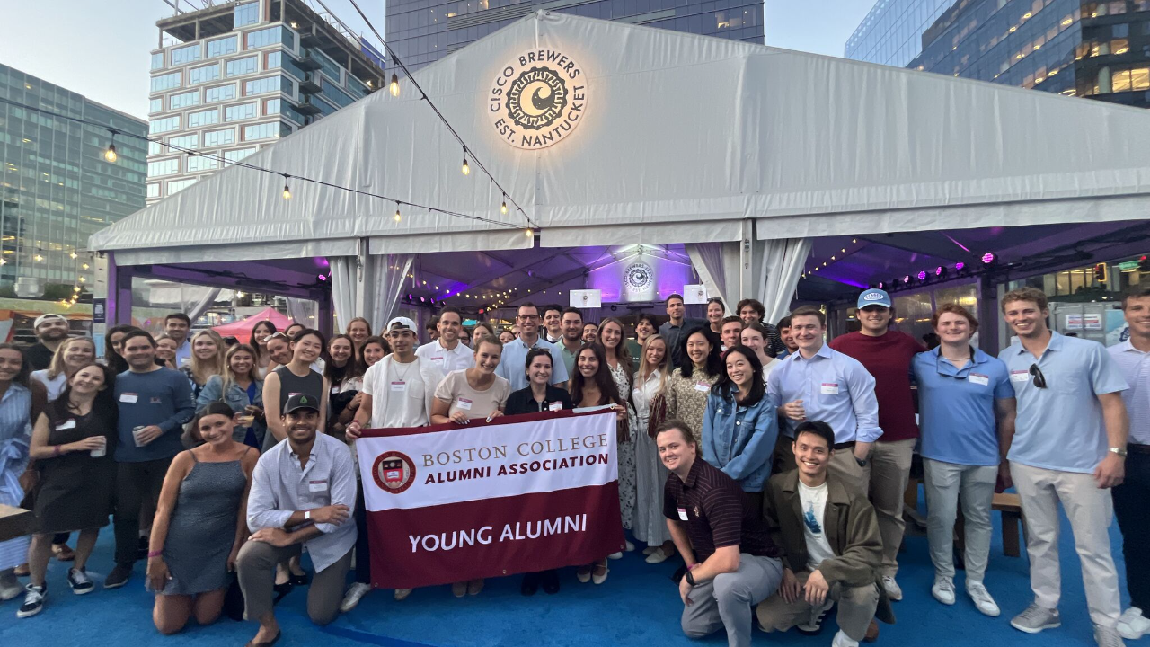 A large group of young adults pose outdoors under a tent, holding a “Boston College Alumni Association Young Alumni” banner, with city buildings in the background.