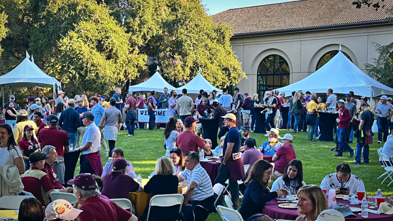 A large group of people gather outdoors on a lawn with white tents, eating, talking, and wearing casual clothing, some in matching team colors. Trees and a building are in the background.