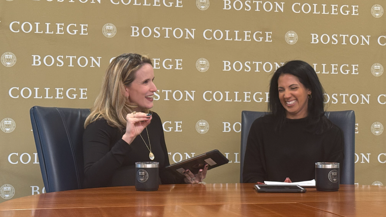 Two women sit and laugh together at a table with Boston College branding behind them. One holds a tablet and gestures while the other smiles, both have mugs and notepads in front of them.