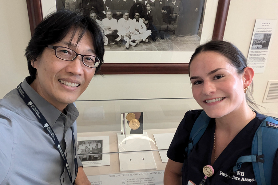 Two people smile for a selfie in front of a glass case displaying a Nobel Prize medal. Behind them is a black-and-white group photo and informational plaques on the wall.