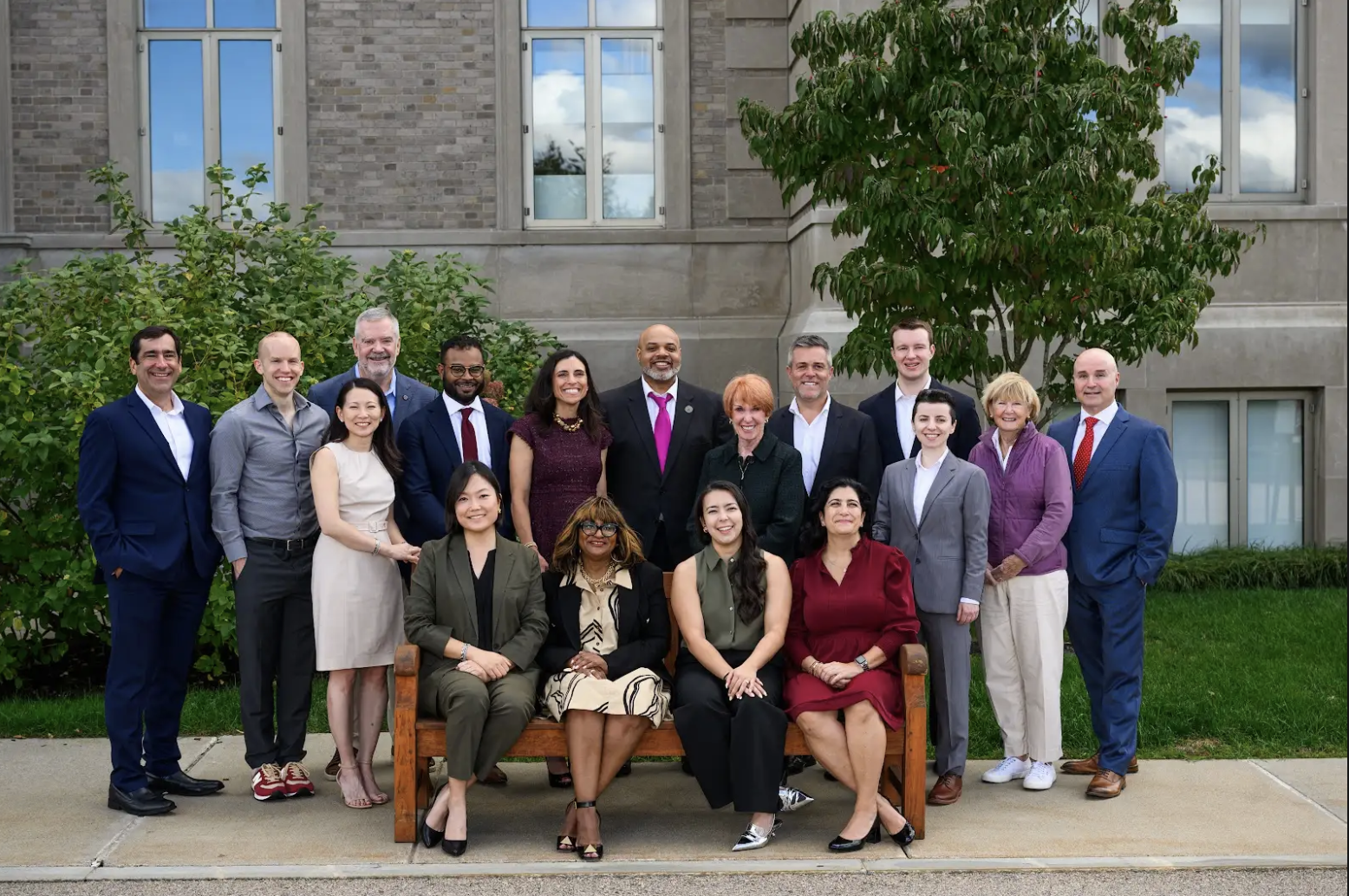 A group of people, diverse in age, gender, and ethnicity, pose together smiling outdoors in front of a building, with some standing and four seated on a wooden bench.