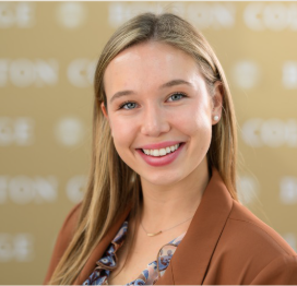 A young woman with long blonde hair, wearing a brown blazer and patterned blouse, smiles in front of a blurred beige background with the word “COLLEGE” partially visible.