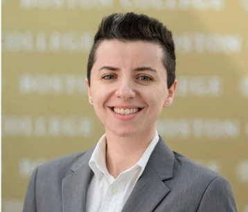 Person with short dark hair in a gray suit and white shirt smiling at the camera, standing in front of a blurred gold background with white text.